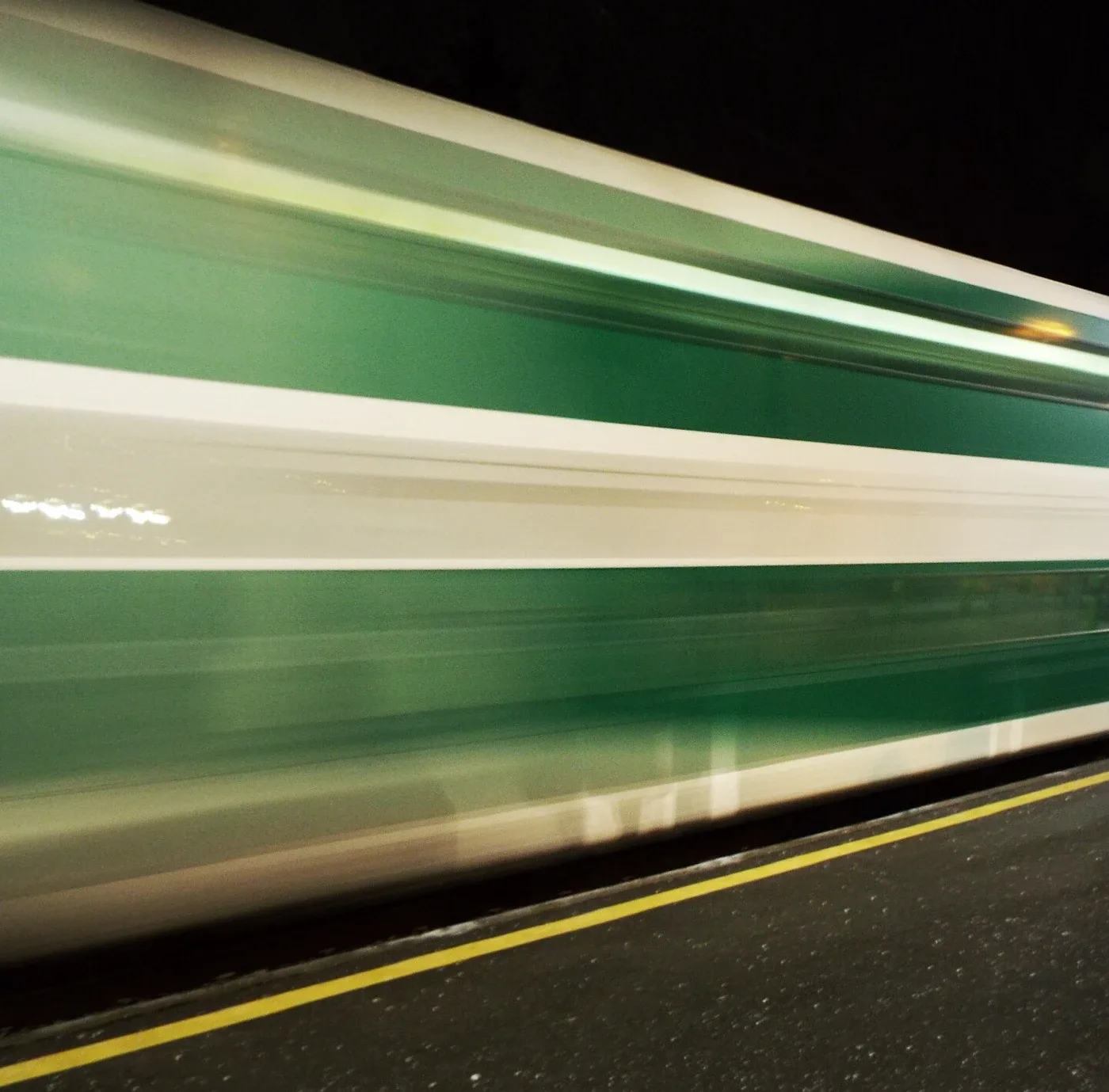 Passing green-and-white train streaks across the frame above a much more clearly defined yellow platform line and speckled platform surface.