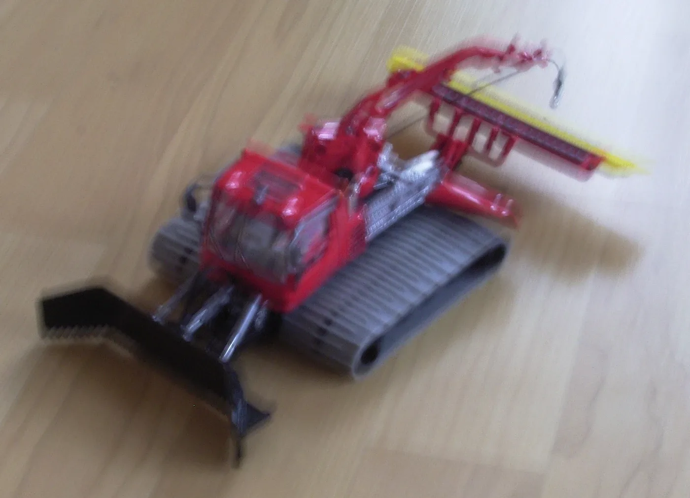 Red model snow groomer and tabletop grain show diagonal smearing, including the cab edges and yellow rear bar.